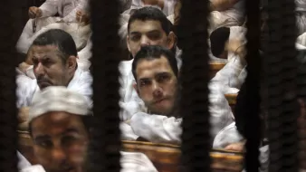 تظهر الصورة مجموعة من الرجال يجلسون في قاعة المحكمة، مع وجود قضبان أو شبك يظهر في المقدمة. يبدو أنهم يرتدون ملابس بيضاء ويظهر على وجوههم تعابير مختلفة. بعضهم ينظر إلى الكاميرا بينما يبدو آخرون مشغولين بأفكارهم. الجو العام يوحي بالتوتر والجدية، مما يعكس طبيعة المكان الذي يصور.