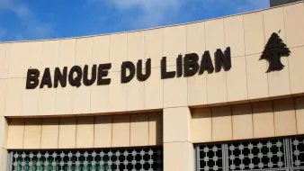 تظهر الصورة واجهة مؤسسة مالية تحمل اسم "بنك لبنان" بالخط العريض. الكتابة باللون الأسود على خلفية فاتحة، وتوجد شجرة الأرز، وهي رمز وطني للبنان، بجانب الاسم. السماء زرقاء مع بعض السحب، مما يعطي انطباعًا بوجود يوم مشمس. يبدو أن الجزئية المعروضة من المبنى حديثة ومرتبة.