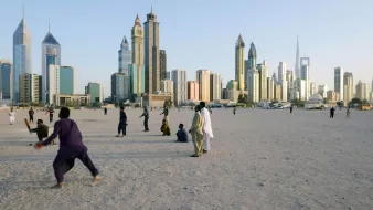 الصورة تُظهر مشهدًا حضريًا لمدينة حديثة، حيث تبرز ناطحات السحاب والمباني الشاهقة. في المقدمة، يوجد مجموعة من الأشخاص يمارسون أنشطة مختلفة على رمال منطقة مفتوحة، ما يوحي بأجواء حيوية. الخلفية تُظهر المدينة مع سماء زرقاء تتخللها أشعة الشمس، مما يضفي لمسة جميلة على المشهد.