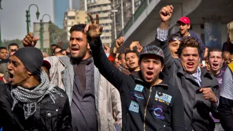 تظهر في الصورة مجموعة من الشباب الذين يقفون في منطقة حضرية. يبدو أنهم يعبرون عن مشاعر قوية، حيث يرفعون قبضاتهم وينظرون إلى الأمام بحماس. تعكس تعابير وجوههم الحماسة أو الغضب، وقد يكونون في حالة احتجاج أو تجمع جماهيري. يحيط بهم مبانٍ عالية وعناصر حضرية، مما يدل على أن النشاط يحدث في مدينة.