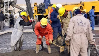 الصورة تظهر مجموعة من العمال في موقع بناء وهم يعملون على صب الخرسانة. يرتدي العمال خوذات لحماية الرأس وأزياؤهم متنوعة الألوان، حيث يركز البعض على تجريف الخرسانة باستخدام معدات يدوية. تظهر في الخلفية شاحنة وأدوات أخرى تتعلق بأعمال البناء، مما يدل على نشاط مكثف في الموقع.