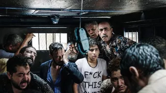 الصورة تظهر مجموعة من الأشخاص الذين يتواجدون في مكان مغلق، يبدو عليهم الارتباك والقلق. هناك مظاهر للفوضى، حيث يظهر بعضهم بمظهر متعب أو مضغوط. الإضاءة في المكان ضعيفة، مما يضيف شعورًا بالتوتر. يبدو أن الوضع في الصورة يعكس حالة صعبة أو حرجة.