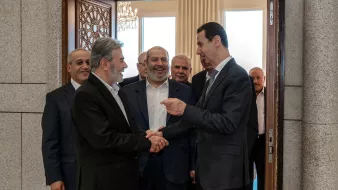 تظهر الصورة مجموعة من الرجال في قاعة استقبال. يتوسط الصورة شخصان يتصافحان، حيث يبدو أن أحدهما في وضعية حديث مع الآخر. الخلفية تحتوي على أبواب وزخرفة رائعة، وهناك أشخاص آخرون يقفون خلفهم، يظهر عليهم تفاعل إيجابي ورغبة في التواصل. الأجواء تبدو رسمية، مما يشير إلى لقاء مهم أو مناسبة خاصة.