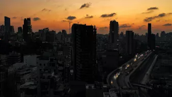تظهر الصورة مشهداً ليلياً لمدينة مكتظة بالمباني والعمارات. السماء تتلون بألوان الغروب، حيث تتدرج الألوان من البرتقالي إلى الأزرق الداكن. في الأسفل، يمكن رؤية طريق يمر بجانب المباني، وقد ظهرت بعض السيارات وهي تسير في الاتجاهات المختلفة. الأضواء بدأت تتلألأ في النوافذ، مما يضفي على المشهد جوًا ساحرًا يعكس حيوية المدينة في المساء.