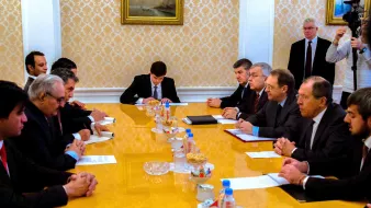 تظهر الصورة اجتماعاً رسمياً يتم فيه نقاش أو تبادل للأفكار بين مجموعة من الأشخاص. يجلس المشاركون حول طاولة كبيرة تحتوي على مستندات وبعض الوجبات الخفيفة. يبدو أن هناك تركيزاً على النقاش، حيث يظهر البعض وهم يكتبون أو يتطلعون إلى المستندات الموجودة أمامهم. الخلفية تحتوي على لوحات فنية تزين الجدران، مما يضيف طابعاً رسمياً إلى الاجتماع.