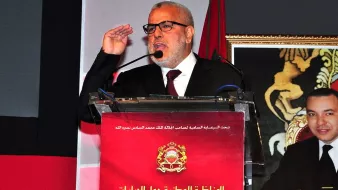 تعذر عليّ التعرف على الشخص أو تقديم تفاصيل محددة حول الصورة. لكن يمكنني وصف ما يمكن أن تحتويه الصورة بناءً على العناصر المألوفة. تظهر الصورة شخصًا يقف أمام منصة تحمل شعارًا رسميًا، ويبدو أنه يتحدث أو يخاطب جمهورًا. الخلفية قد تحتوي على صور أو عناصر دالة على الحدث، مثل العلم أو رمز بلدي. إذا كانت لديك تفاصيل معينة عن الصورة، يمكنني مساعدتك في صياغتها بشكل أفضل.