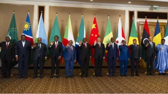 تظهر الصورة مجموعة من القادة أو الممثلين الرسميين من دول مختلفة يقفون في صف واحد. خلفهم، تُعلق أعلام تمثل الدول أو المنظمات المختلفة، بما في ذلك علم الاتحاد الأفريقي. يبدو أن الجميع يلوح بيديه في تعبير عن التحية. يرتدي القادة بدلات رسمية، وبعضهم يرتدي الملابس التقليدية. تعكس الصورة مناسبة رسمية أو قمة تجمع بين الدول.