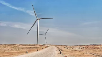 تظهر في هذه الصورة منظرًا طبيعيًا صحراويًا يمتد تحت سماء زرقاء فاتحة. يتواجد في الجزء الأمامي طريق رملي يتجه نحو الأفق، محاطًا بأعمدة صغيرة على جانبيه. في الخلفية، هناك عدة توربينات رياح ضخمة تدور ببطء، مما يُعطي انطباعًا حديثًا حول استخدام الطاقة المتجددة في هذا المناطق القاحلة. يبدو المكان هادئًا وواسعًا، مع تباين واضح بين الألوان الرمادية للأرض والألوان الزرقاء للسماء.