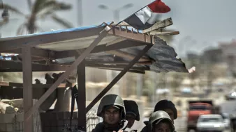 الصورة تُظهر مجموعة من الجنود أو رجال الأمن، يرتدون بدلات عسكرية وخوذ، ويبدو أنهم على أهبة الاستعداد. يقفون خلف حاجز أو نقطة تفتيش، بينما يُمكن رؤية العلم المصري يُرفرف في الخلفية. المشهد يتضمن أيضًا بعض التفاصيل عن البيئة المحيطة، مثل الشوارع والمباني. الأجواء تبدو مشحونة، مما يوحي بوجود حالة تأهب أو تواجد أمني مكثف.