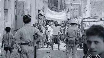 تظهر الصورة مجموعة من الأشخاص في شارع ضيق، يبدو أنهم في خضم حدث أو تظاهرة. في المقدمة، هناك رجل يرفع يديه حاملاً لافتة، بينما يتواجد أفراد من قوات الأمن بالقرب منه. يمكن رؤية الأطفال والبالغين في الخلفية، بعضهم يبدو مهتماً بالحدث. الملابس والإعداد تعكس فترة زمنية معينة، كما يظهر في التفاصيل المعمارية للمنازل. المشهد يحتوي على أجواء من الحماس والتوتر.