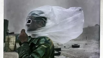 تظهر في الصورة شخص يرتدي زيًا عسكريًا، وقد غطى وجهه بقطع من القماش أو البلاستيك بسبب الظروف الجوية السيئة، مثل العواصف الرملية أو الغبار. يبدو أن الشخص واقف في منطقة غير مستوية، حيث يمكن رؤية بعض الأشياء في الخلفية مثل الإطارات والمواد الأخرى. تعكس الصورة حالة تحدٍ تحملها الشخص في مواجهة البيئة القاسية.