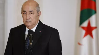 تظهر الصورة شخصية ترتدي بدلة رسمية داكنة، ويقف أمام ميكروفون. في الخلفية، يمكن رؤية علم الجزائر الذي يحمل الألوان الحمراء والبيضاء والخضراء. تعبر تعابير الوجه عن جدية، ويتواجد في مكان يبدو كقاعة رسمية.