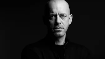 الصورة تُظهر رجلًا ذو رأس أصلع، يقف في ضوء خافت مما يبرز ملامح وجهه بوضوح. عيونه تحمل تعبيرًا جادًا وقويًا، بينما يحدق بشكل مباشر نحو الكاميرا. يرتدي سترة داكنة، ويبدو أن الخلفية سوداء، مما يضيف إلى جمالية الصورة ويعطي إحساسًا بالدراماتيكية.