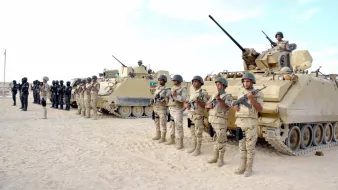 تظهر في الصورة مجموعة من الجنود العسكريين يقفون في صف منتظم، يرتدون زيًا عسكريًا ويحملون أسلحة. خلفهم توجد مدرعات عسكرية، وبعض الجنود يجلسون فوق هذه المدرعات. يبدو أن المشهد قد تم التقاطه في منطقة صحراوية أو رملية، مع سماء صافية في الخلفية. تعكس الصورة أجواءً عسكرية منظمّة وجادة.