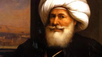 تظهر الصورة رجلاً مسنًّا يرتدي عمامة كبيرة ومجموعة من الملابس التقليدية. يتميز بلحيته البيضاء الكثيفة وابتسامة ودودة. يحمل سيفًا في يده، مما يوحي بمكانته العالية أو دوره القيادي. الخلفية تحتوي على مشهد يبدو كأنه منظر طبيعي لمدينة أو تجمع حضري. تعكس تفاصيل ملابسه وزينه التراث الثقافي لمنطقته.