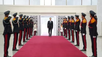 تظهر الصورة مجموعة من الجنود يرتدون الزي العسكري الرسمي، وهم مصطفون على جانبين من ممر مفروش بالسجاد الأحمر. في نهاية الممر، يظهر شخص يرتدي بدلة رسمية يسير نحوهم. الجنود يرفعون أيديهم للتحية، مما يوحي بأن هذه اللحظة تمثل استقبالاً رسمياً أو تكريساً لمناسبة هامة. الخلفية تبدو حديثة مع نوافذ كبيرة تسمح بدخول الضوء.