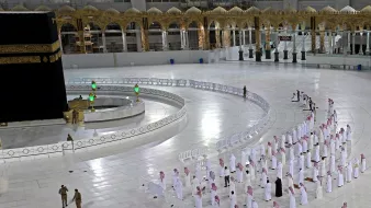 الصورة تُظهر الحرم المكي ومكة المكرمة، حيث يمكن رؤية الكعبة المشرفة في وسط الصورة. هناك عدد من المصلين يقفون في صفوف مرتبة لأداء الصلاة، ويرتدون الملابس التقليدية. الساحة المحيطة بالكعبة نظيفة وواسعة، مع وجود حواجز حول مناطق معينة. كما يظهر بعض رجال الأمن في الصورة، مما يدل على تنظيم المكان. تمثل هذه اللقطة أجواء العبادة والتقرب إلى الله، وخاصة في الأوقات التي تكون فيها أعداد المصلين محدودة.