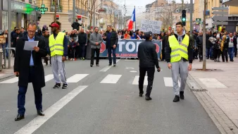 الصورة تظهر مجموعة من الناس يتظاهرون في شارع. في المقدمة، هناك شخصان يرتديان سترة عاكسة، ويبدو أنهما يجريان أو يقومان بمراقبة المظاهرة. في الخلفية، يمكن رؤية لافتة كبيرة مكتوب عليها "STOP ISLAMOPHOBIE"، مما يشير إلى أن المظاهرة تتعلق بمناهضة الإسلاموفوبيا. المتظاهرون يحملون لافتات ويبدو أنهم يتجمعون بشكل منظم. الجو العام يعكس النشاط والحيوية.