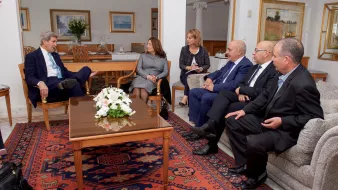 تظهر الصورة مجموعة من الأشخاص يجلسون في غرفة معيشة أنيقة. في المنتصف، يوجد طاولة قهوة تحمل زهورًا بيضاء. يبدو أن هناك نقاشًا أو اجتماعًا يجري بينهم. يجلس شخص يرتدي بدلة زرقاء في الجهة اليسرى، بينما يجلس الآخرون على الأرائك المحيطة. الخلفية تحتوي على لوحات فنية ونباتات، مما يضفي جوًا مريحًا على المكان.