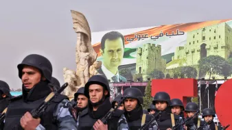 تظهر الصورة عرضًا عسكريًا أو تظاهرة، حيث يسير مجموعة من الجنود في صفوف منظمة، يرتدون زيًا عسكريًا ويحمون أسلحة. خلفهم يوجد لافتة ضخمة تحمل صورة لرجل، تترافق مع مشاهد معمارية تتعلق بسوريا. تظهر أيضًا بعض الرموز الوطنية مثل الأعلام. الأجواء تعكس نوعًا من القوة والتأييد السياسي.