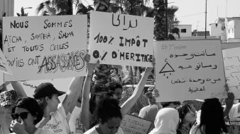 الصورة تظهر مجموعة من الأشخاص يحملون لافتات في مظاهرة. اللافتات تتضمن عبارات تتعلق بالعدالة والحقوق. من بينها نصوص تشير إلى الأحكام الضريبية والمطالبة بالعدالة للضحايا، مما يعكس موقفًا اجتماعيًا أو سياسيًا. الأجواء تبدو حماسية مع حضور العديد من المشاركين.