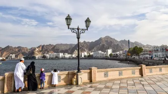 تظهر الصورة منظرًا جميلًا لواجهة بحرية في منطقة جبلية. في المقدمة، يقف شخصان وطفلان على حافة المياه، حيث يستمتع الجميع بالمنظر. الشخصان يرتديان ملابس تقليدية، بينما يرتدي الأطفال ملابس ملونة. في الخلفية، تظهر الجبال الشاهقة ومناظر المدينة مع بعض المباني البيضاء. السماء زرقاء مع غيوم خفيفة، مما يعطي الصورة شعورًا بالهدوء والطبيعة الجميلة. عمود إنارة يضيف لمسة جمالية للمشهد.