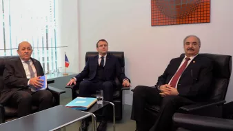 تظهر الصورة ثلاثة رجال يجلسون في غرفة اجتماعات. الشخص في المنتصف يرتدي بدلة داكنة، بينما الأخران يجلسان على جانبيه. يوجد علم فرنسي صغير في الجهة اليسرى من الصورة، والطاولة أمامهم تحتوي على أوراق وزجاج ماء. الجدران محاطة بالزجاج وتحتوي على عمل فني ملون في الأعلى. تعكس التعبيرات والوضوح في الجلسة وجود نقاش أو اجتماعات رسمية.