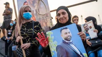 تظهر الصورة مجموعة من الأشخاص في مظاهرة. في المقدمة، هناك سيدة مسنّة ترتدي حجابًا وتمسك بصورة لرجل يبدو أنه قد توفي، بينما تضع يدها الملطخة باللون الأحمر، مما يوحي بمحتوى رمزي. بجانبها، توجد سيدة شابة ترتدي قناع وجه وأمسكت بلافتة. خلفهم، يظهر عدد من الأشخاص الذين يتفاعلون مع الحدث. وتبدو الأجواء مشحونة بالعواطف.