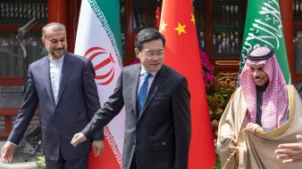 تظهر في الصورة ثلاثة رجال يقفون أمام أعلام ثلاث دول: إيران، الصين، والسعودية. الشخص في الوسط يبدو أنه يتحدث أو يتحرك، بينما الشخصان على الجانبين ينظران إليه. الأجواء تبدو رسمية، وقد تكون الصورة ملتقطة خلال حدث دبلوماسي أو اجتماع. الخلفية تحتوي على نباتات وزينة أخرى تضفي طابعاً احتفائياً.