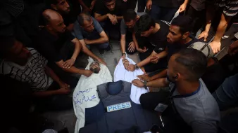A group of people mourns over a deceased individual in a white shroud, surrounded by a press vest.