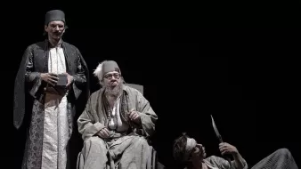 الصورة تظهر مشهداً مسرحياً مع ثلاثة شخصيات. الشخصية الوسطى تجلس على كرسي متحرك، وتبدو في حالة هدوء ورزانة، مرتدية ثياب تقليدية مع عمامة. الشخصيتان الأخريتان تقفان بجانبه، حيث يظهر أحدهما بزي تقليدي أيضاً ويبدو أكثر جدياً، بينما الآخر يظهر بشكل غير مستقر وتمزق في ملابسه، كما يحمل خنجراً. الإضاءة في الخلفية داكنة، مما يبرز الشخصيات في الصورة.