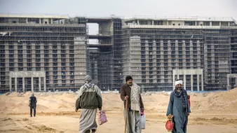 في الصورة نرى مجموعة من الأشخاص يمشون على طريق رملي في منطقة قيد الإنشاء. خلفهم توجد مبانٍ مرتفعة لا تزال في مراحل البناء، مع وجود هياكل معدنية وأخشاب واضحة. يرتدي الأشخاص ملابس تقليدية، ويبدو البعض منهم يحمل حقائب. السماء ملبدة بالغيوم، مما يعطي انطباعًا بجو من الصبر والعمل الجاد في بيئة الإنشاءات.