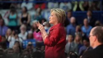 في الصورة، تظهر امرأة ترتدي سترة حمراء وتستخدم ميكروفونًا أثناء إلقاء خطاب أو حديث في تجمع حاشد. خلفها، يوجد جمهور كبير يراقب ويتفاعل مع حديثها. الأجواء تبدو نشطة ومليئة بالطاقة، مع العديد من الأشخاص في الخلفية، مما يدل على أهمية الفعالية.