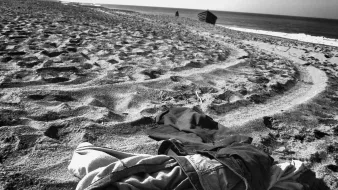 تظهر في الصورة ملابس متكدسة على الرمال على شاطئ. يبدو أن الشاطئ هادئ، حيث تُظهر الأمواج البعيدة خط الأفق. الرمال تحمل آثار أقدام وكأن هناك نشاطًا قد حدث في المنطقة. الأجواء بشكل عام تعكس شعورًا بالوحدة والهدوء.