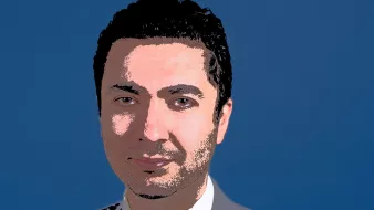 الصورة تظهر شخصاً يرتدي بدلة رسمية باللون الرمادي، مع قميص أبيض وربطة عنق زرقاء. الخلفية ذات لون أزرق، ويبدو على وجهه تعبير واثق. الشعر داكن وممشط بشكل مرتب.