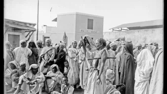 تظهر الصورة مشهداً تاريخياً يضم مجموعة من الأشخاص يرتدون ملابس تقليدية في بيئة حضرية. يتواجد الرجال والنساء بشكل مختلط، بعضهم يجلس وآخرون واقفون، حيث يبدو أنهم يعيشون لحظة تتسم بالنقاش أو الاحتجاج. الخلفية تضم مباني بسيطة، مما يشير إلى بيئة قد تكون في منطقة شمال إفريقيا أو الشرق الأوسط. تعكس الملابس والتعبيرات الثقافية تفاصيل الحياة الاجتماعية في تلك الفترة.