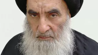 الصورة تُظهر رجلًا مسنًّا ذو لحية بيضاء وشعر داكن تحت عمامة سوداء. يبدو عليه الوقار والجدية. يرتدي ثوبًا داكنًا، مما يعكس نمطًا تقليديًا في اللباس. الخلفية بسيطة ووردية اللون، مما يبرز ملامح وجهه.