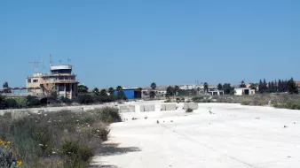 الصورة تظهر مشهداً لمدرج مهجور أو موقع كان مخصصاً للطيران. في الخلفية، يمكن رؤية برج مراقبة وبعض المباني التي تبدو قديمة ومهجورة. الأرضية بيضاء وهي عبارة عن سطح خرساني، وهناك بعض الأعشاب والنباتات الصغيرة تنمو حولها. السماء واضحة وزرقاء، مما يضفي شعوراً بالمسافة والهدوء.