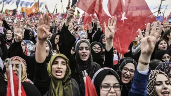 الصورة تظهر حشودًا من النساء في تظاهرة أو فعالية جماهيرية. العديد من النساء يحملن الأعلام التركية ويعبرن عن حماسهن من خلال رفع الأيادي. يبدو أنهن يرتدين ملابس تقليدية أو حجاب، مما يعكس تنوع أساليب التعبير عن الهوية. الجو العام يظهر حيوية وشغفًا، مع وجود لافتات وأعلام في الخلفية تدل على رسالة أو هدف مشترك.