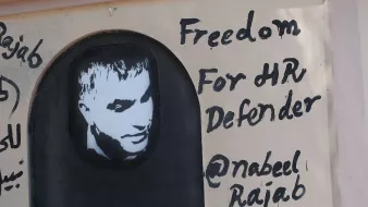 الصورة تظهر جدارية على حائط، تحتوي على صورة لشخص يبدو وكأنه إنسان. تحت الصورة، هناك نص مكتوب باللغة الإنجليزية يقول "حرية لمدافع حقوق الإنسان" مع ذكر اسم "@nabeel Rajab". الألوان المستخدمة تتضمن الأسود والأبيض، حيث تبرز الصورة بشكل يلفت الانتباه. تعبر الجدارية عن دعم لشخصية معروفة في مجال حقوق الإنسان.
