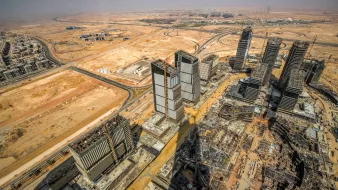 تظهر الصورة منظرًا جويًا لمدينة قيد الإنشاء في منطقة صحراوية. يمكن ملاحظة العديد من الأبراج والمباني الحديثة التي تُشيد، بالإضافة إلى الطرق الواسعة التي تلتف حول المشروع. يحيط بالمكان أرض جرداء، مما يبرز التباين بين البناء الحديث والطبيعة القاحلة. كما تُظهر الصورة التطور العمراني السريع في تلك المنطقة.