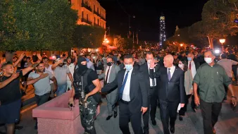 تظهر الصورة مجموعة من الأشخاص في شوارع مدينة مضاءة بأضواء ليلية. يتواجد في المقدمة رجل يرتدي بدلة مع أشخاص آخرين، من بينهم رجل ملثم يحمل سلاحًا. يبدو أن هناك تجمعًا من الحشود في الخلفية، حيث يتفاعل بعضهم مع الموقف. الأجواء توحي بوجود حدث أو مظاهرة، ومشاعر الحماس والانفعال واضحة في تعابير الحضور.