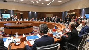 تظهر الصورة اجتماعًا يُعقَد في قاعة مؤتمرات، حيث يجلس العديد من المشاركين حول طاولة دائرية. تتواجد شاشات عرض أمام بعض الأشخاص، وتبدو القاعة مزينة ببعض الزهور. يُلاحظ وجود عدد من الحضور في الخلفية، مما يشير إلى أهمية الحدث. المشاركون يرتدون ملابس رسمية ويظهرون في لحظة من النقاش أو الاستماع.