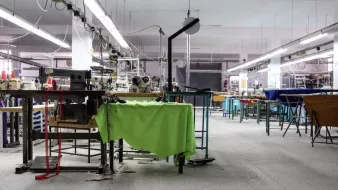 تظهر الصورة مشهدًا داخليًا لورشة خياطة. المساحة تبدو واسعة ومشرقة، حيث توجد آلات خياطة متعددة منتشرة في المكان. بعض الطاولات تحتوي على أقمشة ملونة، في حين أن العديد من المعدات والأدوات موجودة في حالة استخدام أو جاهزية. الأرضية مغطاة ببعض القصاصات من القماش، مما يشير إلى نشاط العمل. الإضاءة تأتي من الأنوار السقفية، وهناك ساعة على الحائط تدل على الوقت. يبدو أن الورشة تحتاج إلى بعض الترتيب.