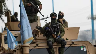 تظهر في الصورة مجموعة من الجنود العسكريين أثناء وجودهم في مركبة مدرعة. يرتدي الجنود زيًا عسكريًا ويظهر على بعضهم معدات تكتيكية وأسلحة. أحد الجنود يجلس على الجزء العلوي من المركبة، بينما يظهر آخرون يقفون عليها. هناك علم أزرق يعود لجهة معينة يبدو أنه متعلق بمهام حفظ السلام. الأجواء المحيطة تبدو حضرية مع وجود أشجار ومرافق عامة في الخلفية.