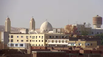 الصورة تظهر مجموعة من المباني في منطقة حضرية، حيث يتواجد مباني ذات طراز معماري مميز، بما في ذلك الكنائس التي تتضمن قبابًا وجرسًا. الخلفية تحتوي على تلال وصحراء، مما يوحي بأن المنطقة قد تكون قريبة من الطبيعة. يوجد أيضًا مجموعة من المباني السكنية المنخفضة، تظهر على اليمين، مما يعطي انطباعًا عن الحياة اليومية في تلك المنطقة.