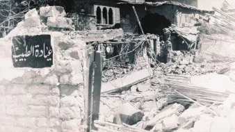 الصورة تُظهر مشهداً لمدمرات بسبب أحداث سابقة، حيث يظهر حطام المباني والخرائب. في مقدمة الصورة، توجد لافتة تحمل اسم مكان، ولكنها مهملة وسط الأنقاض. الخلفية تحتوي على نوافذ وأجزاء من مباني قديمة، مما يعكس تأثير الزمن أو كارثة معينة. الأجواء توحي بالحزن والألم الناتج عن الدمار.