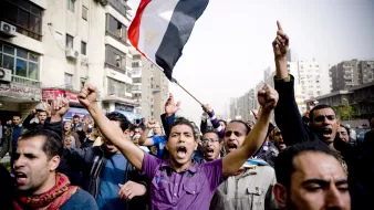 الصورة تظهر مجموعة من الناس في مظاهرة حاشدة. في المقدمة، يوجد رجل يحمل علم مصر ويبدو أنه يرفع صوته عالياً تعبيراً عن الحماس أو المطالب. خلفه يظهر حشد كبير من المتظاهرين، بعضهم يرفعون أيديهم ويهتفون. تعكس الصورة أجواء من النشاط والمشاركة الشعبية، مع مباني المدينة في الخلفية تحت سماء واضحة.