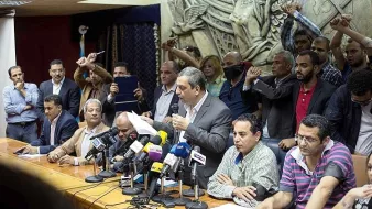 تظهر الصورة اجتماعًا تضامنياً أو مؤتمرًا صحفيًا حيث يجلس عدد من الأشخاص حول طاولة كبيرة، بينما يقف متحدث أمامهم يتحدث إلى الحضور. في الخلفية، يرفع آخرون أيديهم في إشارة إلى الاحتجاج أو التأييد. يظهر البعض في الصورة تفاعلهم مع المتحدث، مما يشير إلى أجواء حماسية. الخلفية تحتوي على منحوتات أو أنماط تزيينية، مما يضيف لمسة رسمية لمكان الاجتماع.