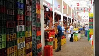 تظهر الصورة سوقاً تقليديًا مزدحمًا، حيث تتواجد صفوف من الأكشاك والمتاجر تحت سقف مغطى. يوجد العديد من الأشخاص يمارسون التسوق، ويتجولون بين الخضروات والفواكه المُعروضة. الجدران محاطة بصناديق ملونة مصنوعة من البلاستيك، مما يضفي جواً حيوياً على المكان. في الخلفية، يمكن رؤية الأعلام التونسية معلقة تُعبر عن الهوية المحلية.